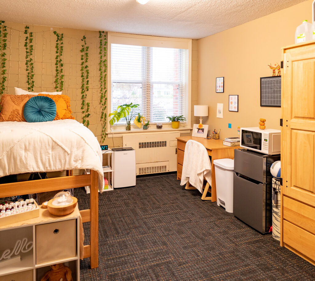 Interior of Hedgpeth Hall room including a twin bed, decorative ivy hanging from the wall, a desk, mini fridge, microwave and wardrobe.